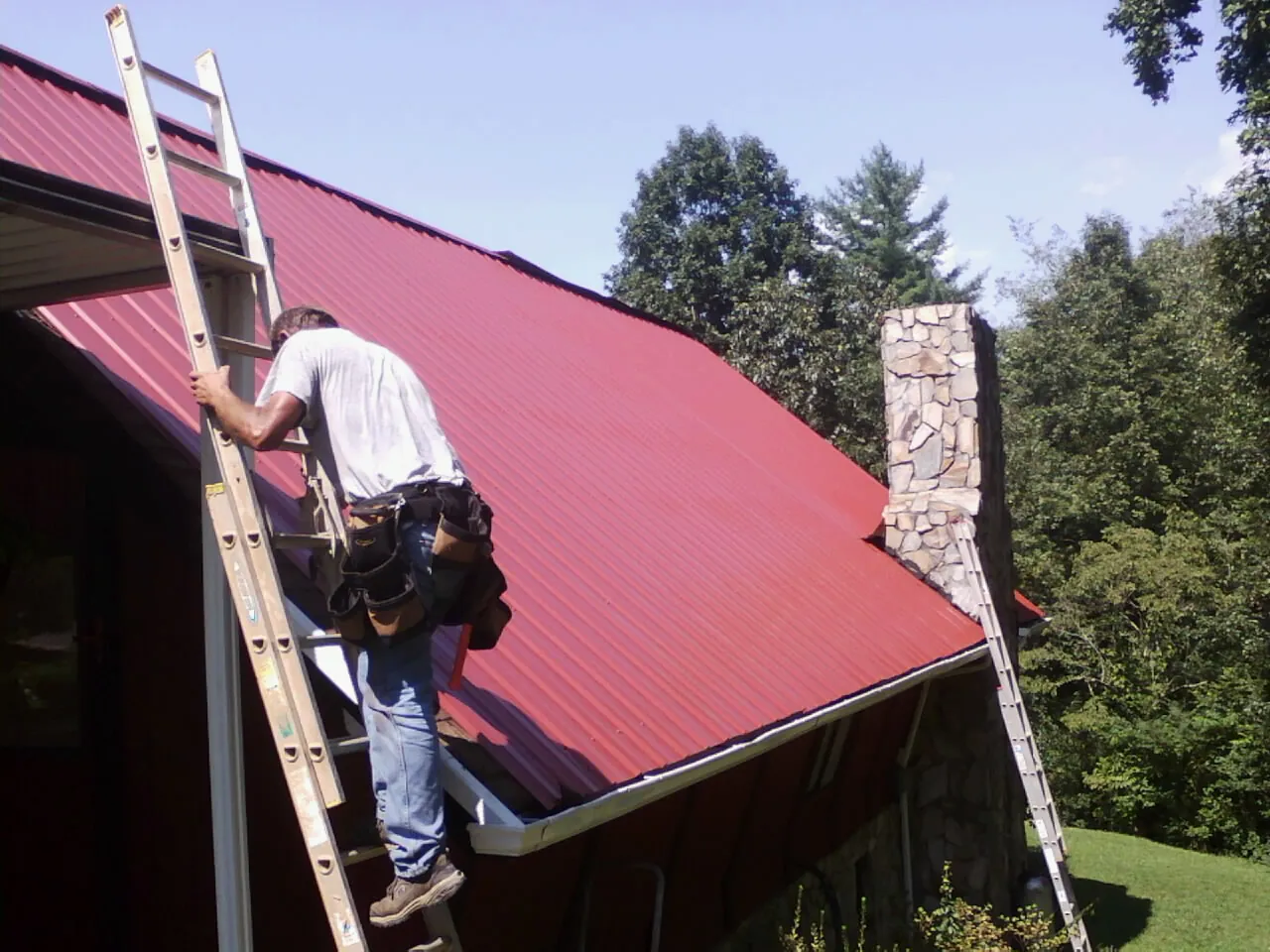 Roofer on a ladder beside a red metal roof and stone chimney — Soffit & Fascia Repair by Roofing Brothers in Minturn